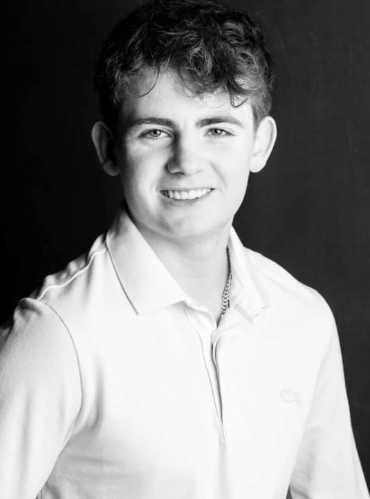 A black and white portrait of a smiling young man with wavy hair, wearing a light coloured collared shirt.